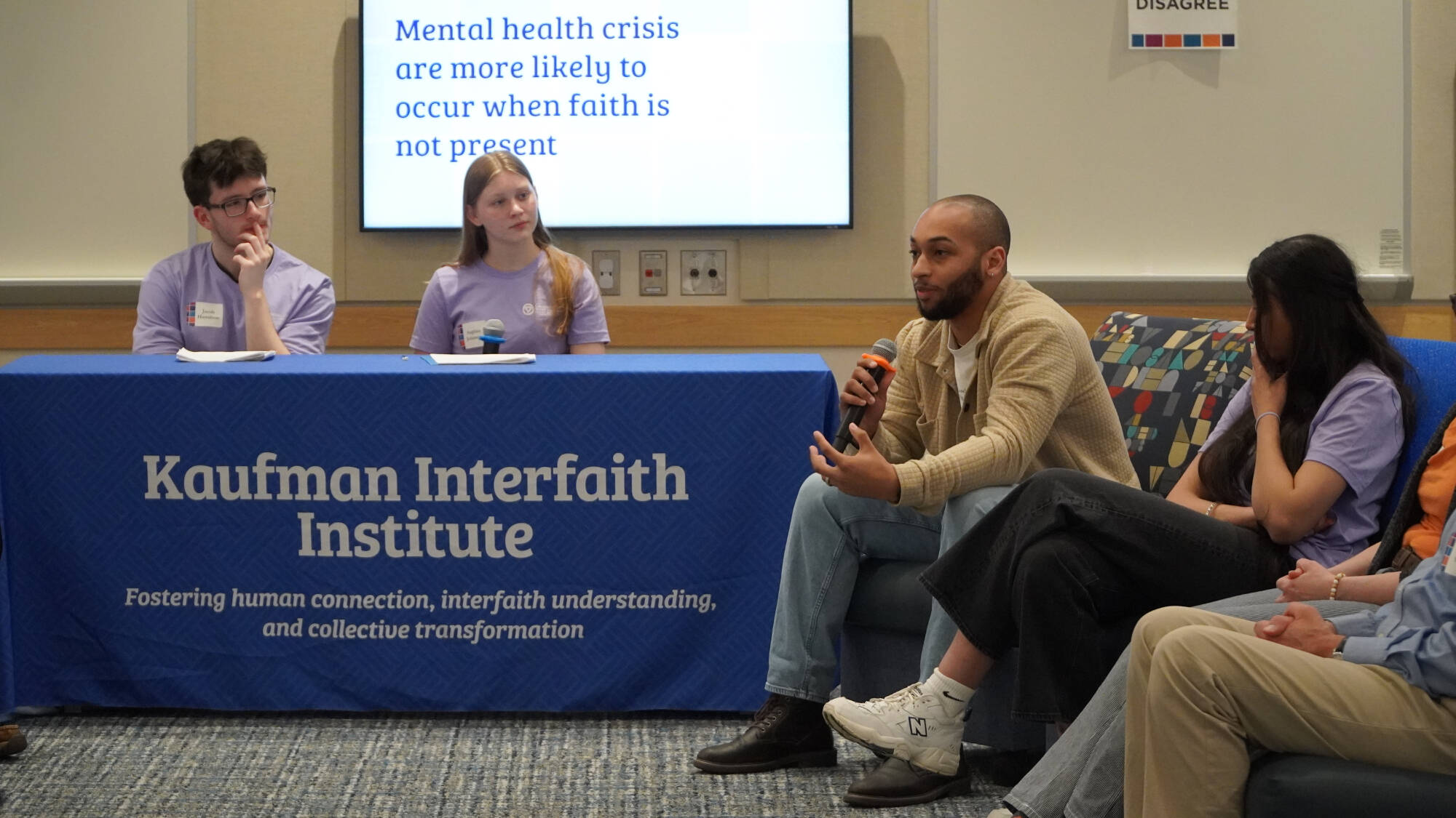 Two Scholars sitting at a table with the Kaufman banner listening to a seated man speak into a microphone